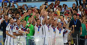 View of the celebrating German team on a podium behind a glass screen, Lahm holding the trophy aloft, while spectators with cameras stand behind