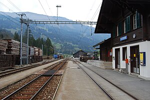Zweistöckiges Gebäude mit Satteldach neben drei Eisenbahnschienen und zwei Bahnsteigen
