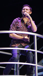 A young man wearing a check shirt and jeans, singing into a microphone