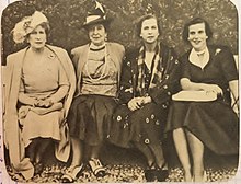 čtyři dámy sedící na lavičce, Victoria-Eugenie ve světlých šatech s pernatým kloboukem, Henriette ve světlých šatech s malým kloboukem, Marie-José v tmavých šatech se světlými vzory a Lilian, usmívající se v tmavých šatech a v ruce drží knihu klín