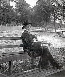 Foto van een oudere man in een pak en met een stok zittend op een bankje in een park