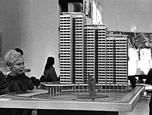 Blonde Frau links im Bild, betrachtet ein Modell eines Hochhauses