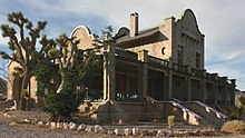 An ornate, two-story masonry building rests under a partly cloudy sky in a setting of gravel, two spiny trees, and many low shrubs. Its windows are boarded.