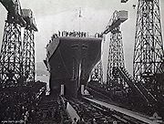 Photograph of the bow of an aircraft carrier as it slides backwards down a slipway. Crowds are gathered around the slipway, underneath several cranes