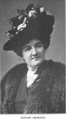 A white woman with dark hair and eyes, wearing a dark hat with abundant embellishments, and a fur wrap