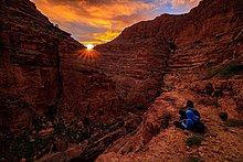 Tørre landformer.  To mennesker ser solen forsvinne bak fjellet og sitter på et odde over en dal som du ikke kan se bunnen av.