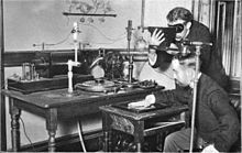 Using early Crookes tube X-Ray apparatus in 1896. One man is viewing his hand with a fluoroscope to optimise tube emissions, the other has his head close to the tube. No precautions are being taken. Crookes tube xray experiment.jpg