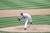 Un homme vêtu d'un uniforme de baseball blanc et d'une casquette de baseball bleue lance une balle de baseball depuis un monticule de terre sur un terrain en herbe.  Il lance avec sa main gauche, et il y a un gant de baseball noir sur sa main droite.