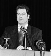 Photo en noir et blanc de John Travolta en costume cravate et assis derrière une table avec un micro devant lui.