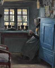 En gammel strikkende kone fra Lille Næstved, set fra venstre side, siddende ved vinduet i en stue, i hvilken et blåmalet skab til højre i forgrunden