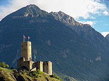 Fotografia colorida de um castelo fortificado em um afloramento rochoso, torre de menagem cilíndrica e montanha ao fundo.