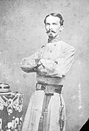 Black and white photo shows a man with a moustache and Van Dyke beard. His arms are folded and he wears a gray military uniform.