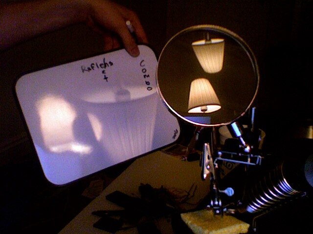 Real image of a lamp is projected onto a screen (inverted). Reflections of the lamp from both surfaces of the biconvex lens are visible.