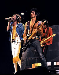 The Rolling Stones di atas panggung pada bulan Desember 1981. Dari kiri: Mick Jagger mengenakan jaket biru dengan pakaian kuning dan ikat pinggang hitam bernyanyi di depan mikrofon, Keith Richards mengenakan celana hitam dan rompi ungu kecil (tanpa kemeja) memainkan gitar hitam diiringi kiri—dan sedikit di depan—Jagger, Ronnie Wood mengenakan jaket oranye dan kemeja/celana hitam memainkan gitar krem ​​​​di belakang Jagger dan Richards.