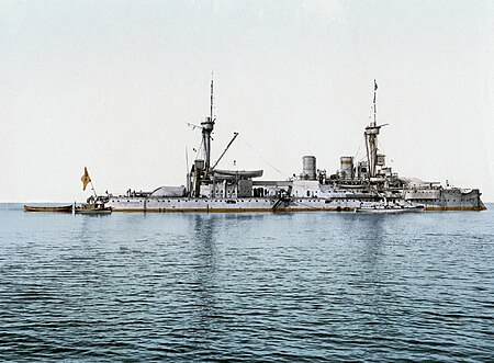 A large gray battleship with two tall masts sits idly in calm waters. Three small boats are tied alongside.