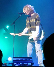 um homem vestido e com cabelo como Kurt Cobain tocando guitarra no palco.