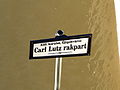 Photographie d'un panneau signalétique indiquant en hongrois, en lettres noires sur fond bland "XIII. kerület, Újlipótváros" et "Carl Lutz rakpart".
