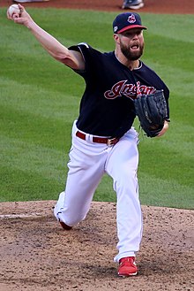 A man in a dark blue baseball jersey with the Cleveland Indians' text logo on the front and dark blue cap with the Chief Wahoo logo delivers a pitch with his right hand.
