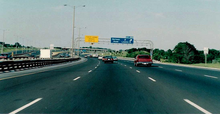 "Driving down a wide highway during the day. Infront are two signs mounted on an overhead lattice. The left sign marks the upcoming end of a lane. In the distance, several highway lanes converge, leaving a large dirt median in the centre."