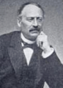 head-and-shoulders of man with moustache, hand on chin and wearing dark jacket, waistcoat, white shirt and bow tie