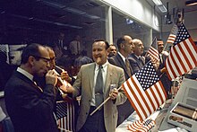 Fotografia a colori di Kraft e altri funzionari, incluso Gilruth, che si congratula con se stessi per il primo sbarco sulla luna di successo accendendo sigari e tenendo in mano bandiere americane.