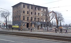 Fotografia barwna, na pierwszym planie torowisko tramwajowe, za nim jezdnia i chodnik, po którym idą piesi, na nim z lewej murowany czterokondygnacyjny budynek podczas rozbiórki za pomocą żółtej maszyny, która znajduje się na prawo od budynku