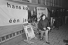 Black and white photograph of people standing on street in front of banner