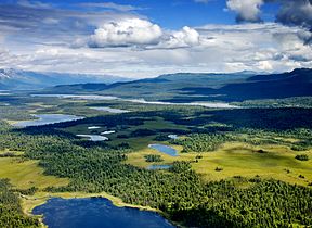 Aerial photograph of several lakes, interspersed with conifer forests and meadows, with tall mountains in the distance and clouds and blue sky overhead. The clouds are casting shadows over the forests and lakes.