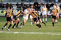 Image shows a rugby league match being played between two teams. A player from the attacking team is about to be tackled by three members from the defensive team