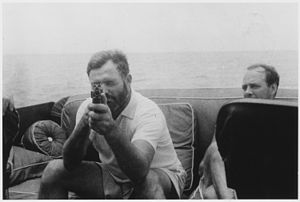 A bearded man sitting on cushions on a boat in the sea aims a gun at the camera; the man looking next to him looks exasperated.