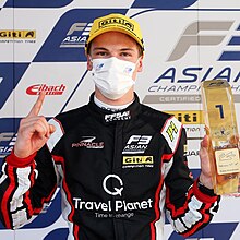 Fotografia a colori di un giovane uomo, con una tuta da pilota nera, maschera bianca, che tiene in una mano il trofeo del vincitore.