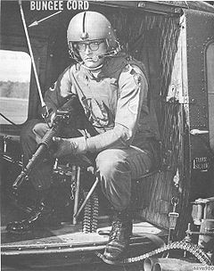 A U.S. Army Vietnam-era door gunner (c. 1966) is shown manning his duty position on a UH-1B/C helicopter gunship, with a 'bungee cord' securing his M60 machine gun to the aircraft cabin doorway.