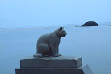 Fotografía en color (color dominante = azul) de una escultura de piedra de un gato sentado, visto de perfil.  Un cuerpo de agua al fondo.