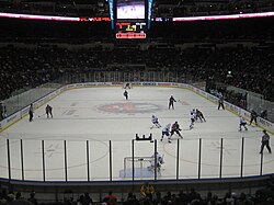 An ice hockey rink on which two teams are playing seen from some distance up and behind the goal. High transparent shielding mounted on the wall surrounds the rink.