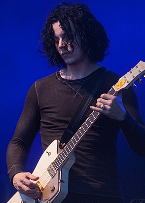 A photo of a caucasian man dressed in a brown thermal-knit shirt performing with a white guitar.