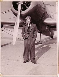 Black and white photograph of Mollison taken in 1936. He is looking into the camera, wearing a suit, shirt, tie and flying helmet. He is standing in front of an aeroplane near its propeller.
