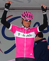 Un uomo con un casco rosa che indossa un top rosa con scaldabraccia e guanti neri.  Ha le mani sopra la testa