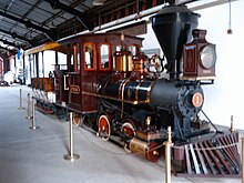 Uma locomotiva a vapor vermelha com um arranjo de rodas 0-4-2T (sem rodas dianteiras, quatro rodas motrizes e duas rodas traseiras) e sem tender, acoplado a um pequeno vagão