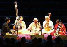 Eine Gruppe von fünf Männern und Frauen sitzt auf einer Plattform, zwei spielen langhalsige Lauten, zwei andere spielen Streichinstrumente und einer stützt seine Hände neben Trommeln auf.