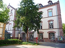 Edificio a due piani intonacato rosa molto chiaro con rivestimenti in arenaria rosa.  In primo piano un grande albero alto quanto l'edificio.