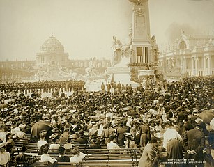 Фотография в сепии, изображающая большую толпу, собравшуюся перед колонной с американским флагом.