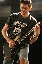 un homme avec un tshirt noir et un jean troué jouant de la guitare.