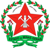 A coat of arms with a large red star in the middle bearing mining equipment. Above the star are the words "Libertas Quae Sera Tamen." The coat of arms is surrounded by a wreath of red berries and green leaves, with two stems of leaves sprouting from the bottom and framing a banner stating "Estado de Minas Gerais." A green ribbon is at the bottom; on the left tail, it says "15 de junho" and on the right, "de 1891."