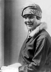A young smiling woman wearing an embroidered hat and a jacket with furred collar and sleeve hems.