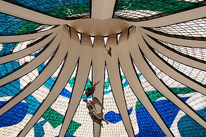 Interior da Catedral Metropolitana de Brasília, Brasil. Desenhada por Oscar Niemeyer e projetada por Joaquim Cardozo. Concluída e consagrada em 31 de maio de 1970, a catedral é uma estrutura hiperboloide construída com 16 colunas de concreto, pesando 90 toneladas cada. Na praça de acesso à catedral estão quatro esculturas de bronze de 2,5 metros de altura representando os quatro Evangelistas, criadas pelos escultores Alfredo Ceschiatti e Dante Croce em 1968. Também fora da catedral, à direita, como visitantes de frente para a entrada, ergue-se um campanário de 20 metros de altura contendo quatro grandes sinos doados por espanhóis residentes no Brasil e fundidos em Miranda de Ebro. Na entrada da catedral há um pilar com passagens da vida de Maria, mãe de Jesus, pintadas por Athos Bulcão. (definição 3 715 × 2 468)