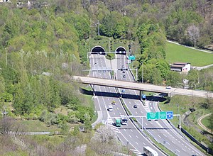 A highway road with two lanes in one direction and three lanes in the other one, merging into two separate tunnels. The highway is surrounded by green forest.