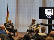 A crowd listens as a staff member from the German Embassy and Eric Jarosinski, a. k. a. Nein Quarterly, sit in modernist chairs on a low stage. There is a black, red and yellow German flag behind Jarosinski and a flat-screen behind the embassy person displaying the Nein Quarterly avatar. A diffuse light through high curtains creates a blue-grey neutral far background.