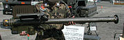 A large pipe-like weapon with a box on its lower lift hand side and upper left hand side, held up for display to the camera. A sign written in Japanese and a military truck can be seen in the background.