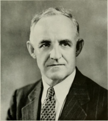 A black and white photograph of a white man in a suit, shot from the chest up