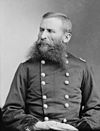 Man with dark dual-pointed moustache sitting down facing left with hands crossed in uniform with two columns of buttons